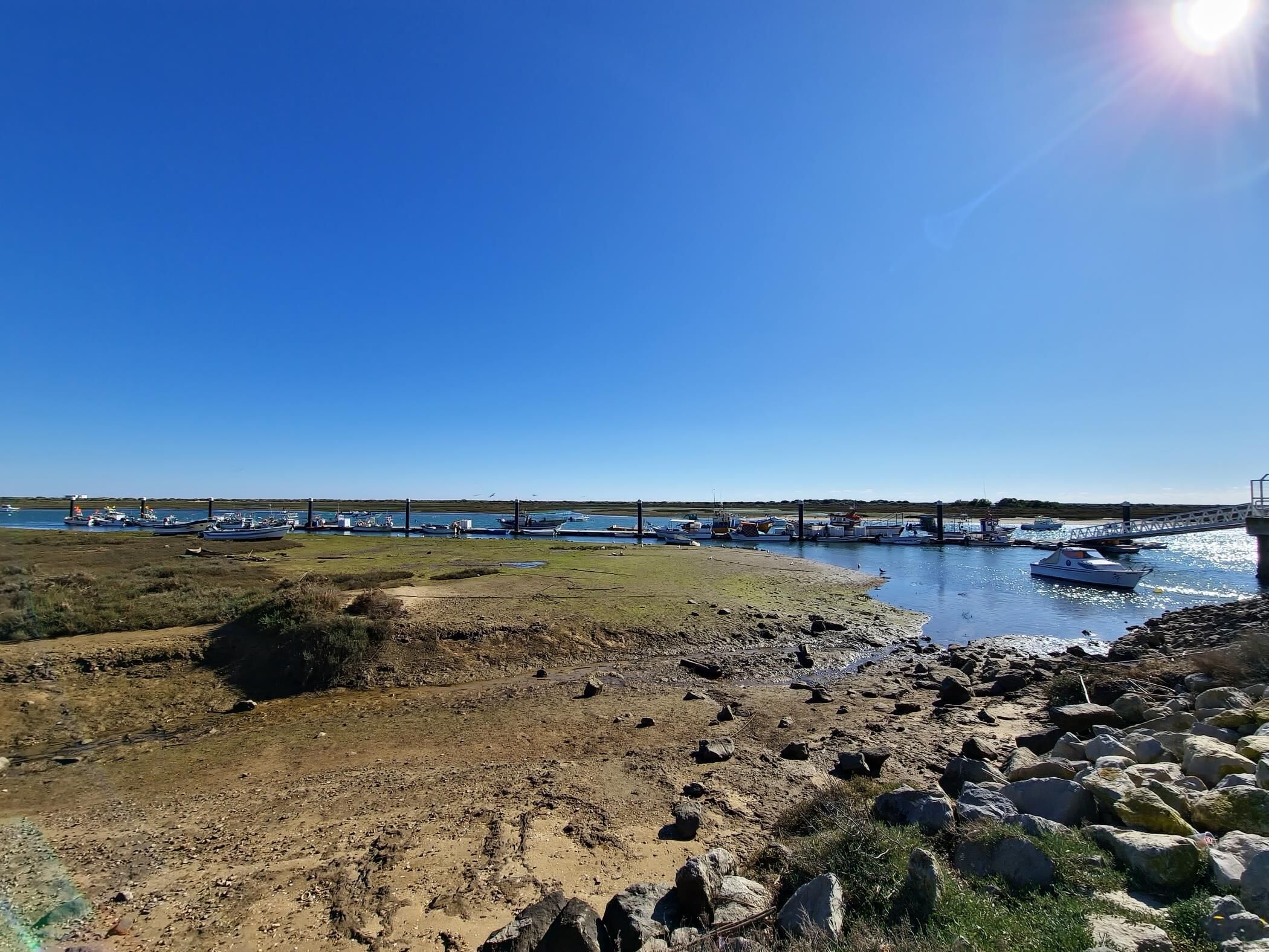 A coastal landscape with boats docked at a pier, rocky terrain, and a clear sky.