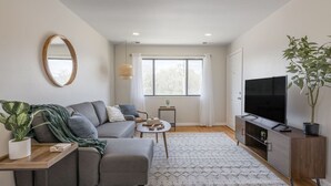 A modern living room with a grey sofa, a coffee table, a TV, and a potted plant. - Furnished 2BR Retreat with Private Garage (Kansas City)