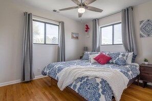 Un dormitorio con cama, ventilador de techo, dos ventanas con cortinas, mesita de noche y una planta colgada en la pared.