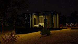 A modern, glass-walled cabin with a bed and a guitar, surrounded by trees and a chain-link fence.