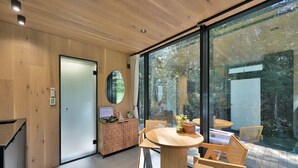 A modern interior with a wooden reception desk, a round wooden table with chairs, and large glass windows offering a view of trees.