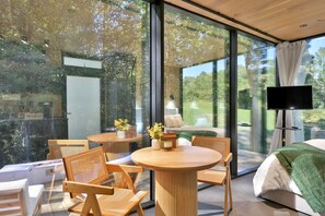 A modern room with large glass windows, a round wooden table, wicker chairs, a bed with a green blanket, and a TV on a stand. - ÖÖD Texas Mirror House (Houston) (Coldspring)