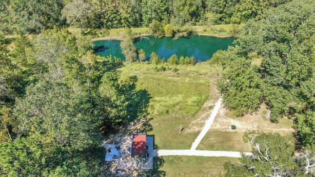 Vue aérienne d’une propriété dotée d’un grand étang, entourée d’une forêt dense. On y aperçoit un petit bâtiment avec un toit rouge et un chemin qui y mène.