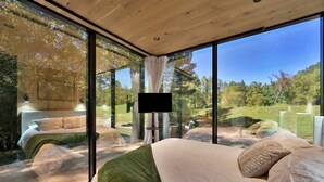 Une chambre moderne dotée d’un grand lit, d’une télévision et offrant une vue sur une zone boisée grâce à de grandes fenêtres.