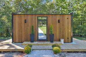 A wooden cabin with a door, two potted plants, a small porch, and a gravel path leading to a wooden deck. - ÖÖD Texas Mirror House (Houston) (Coldspring)