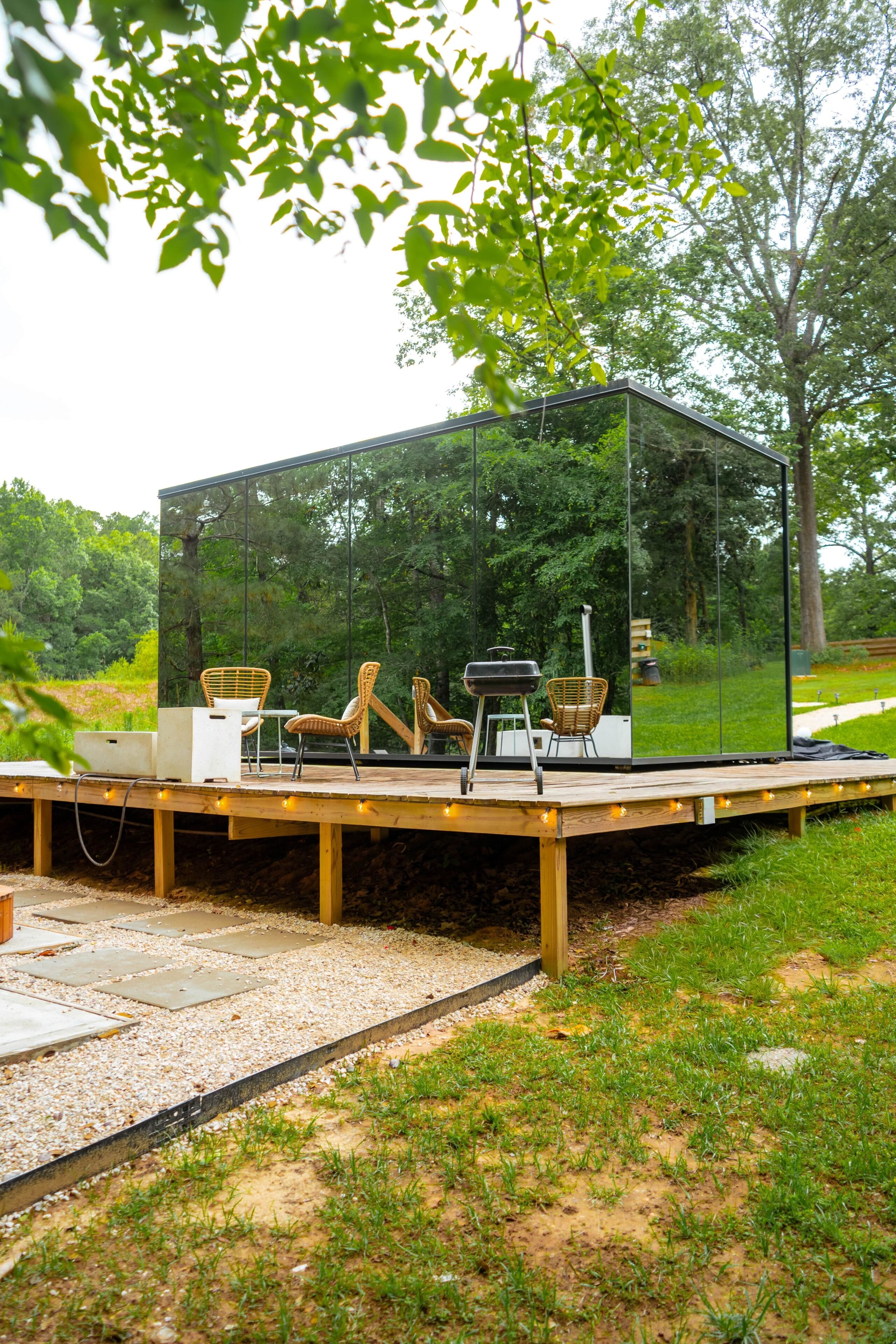A transparent glass structure with outdoor furniture, surrounded by greenery.