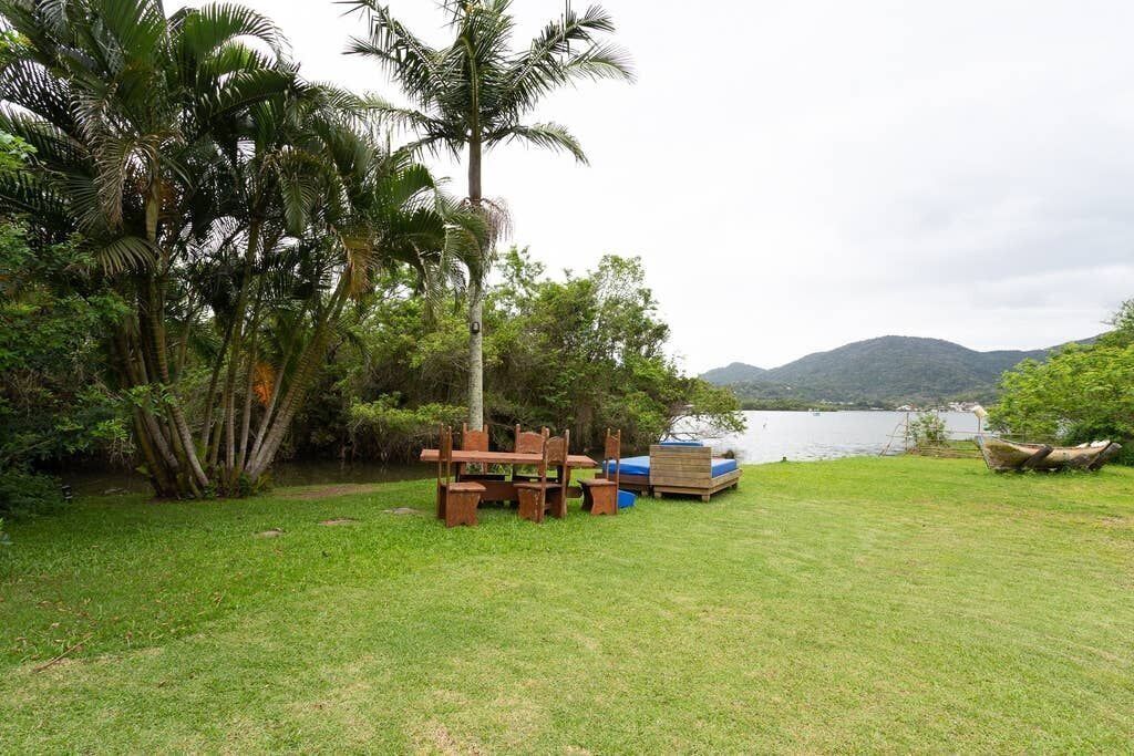 A grassy area with a wooden table and chairs, a blue mattress, and a boat near a lake with mountains in the background.