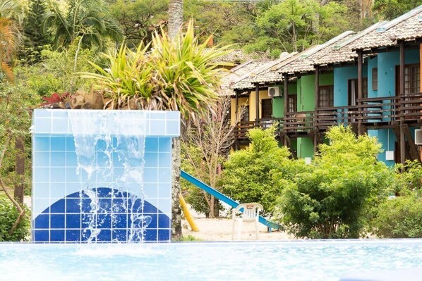 Una piscina con cascada, tobogán y un edificio con balcón al fondo.