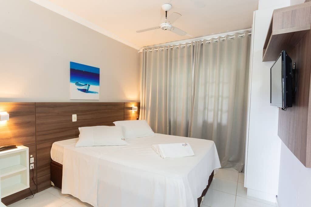 Habitación de hotel con cama, ventilador de techo, un cuadro en la pared, televisión montada y una ventana con cortinas.