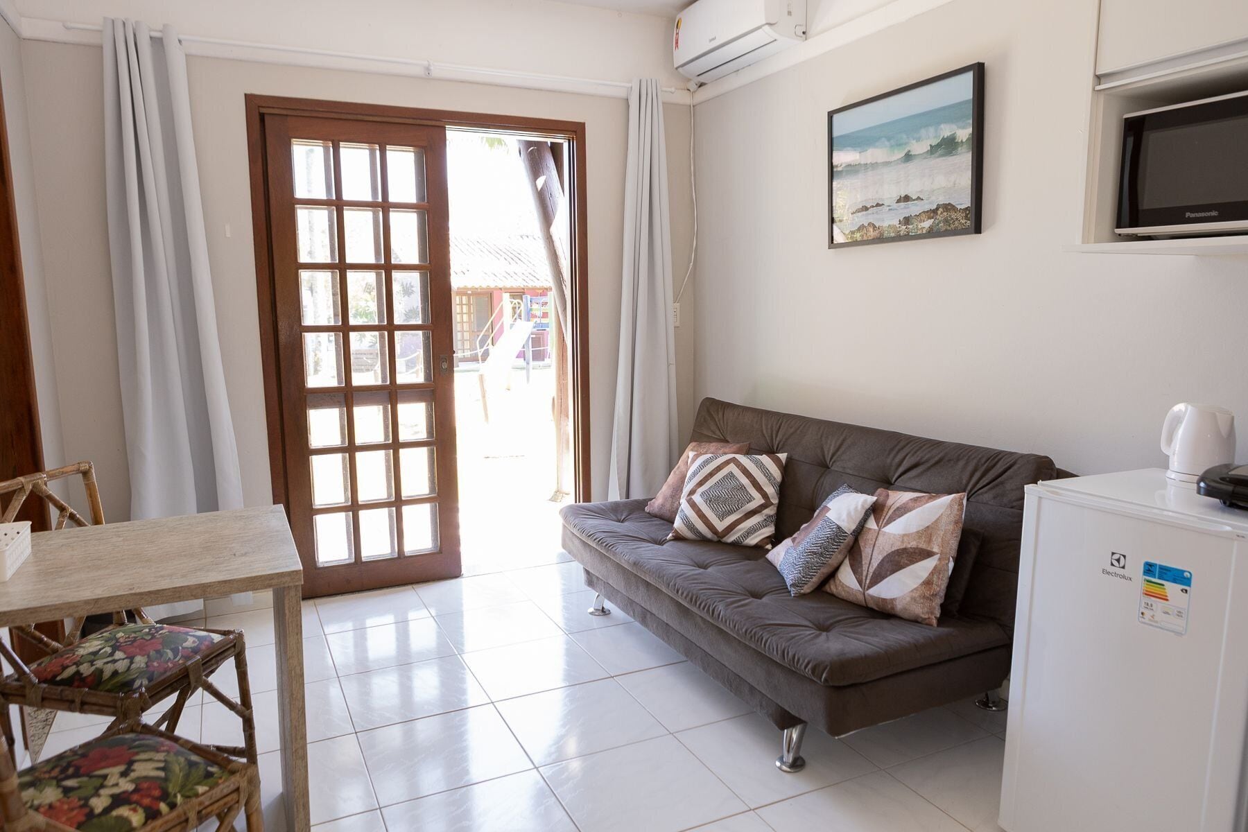 A living room with a sofa, a small table, and a chair. There is a door leading outside, a wall-mounted TV, and a refrigerator.