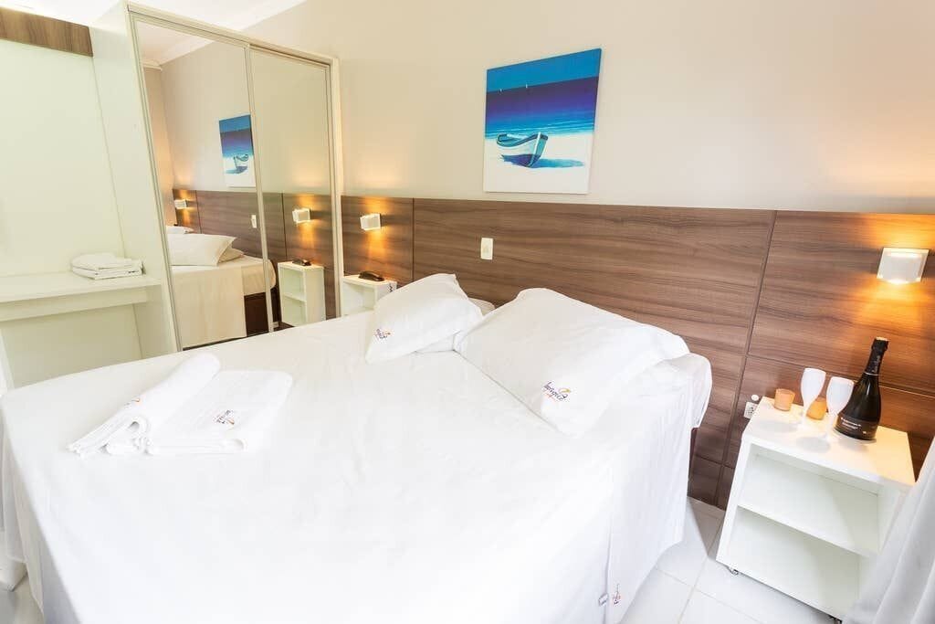 A hotel room with a large bed, a bedside table with a bottle and glasses, a mirror, and artwork on the wall.