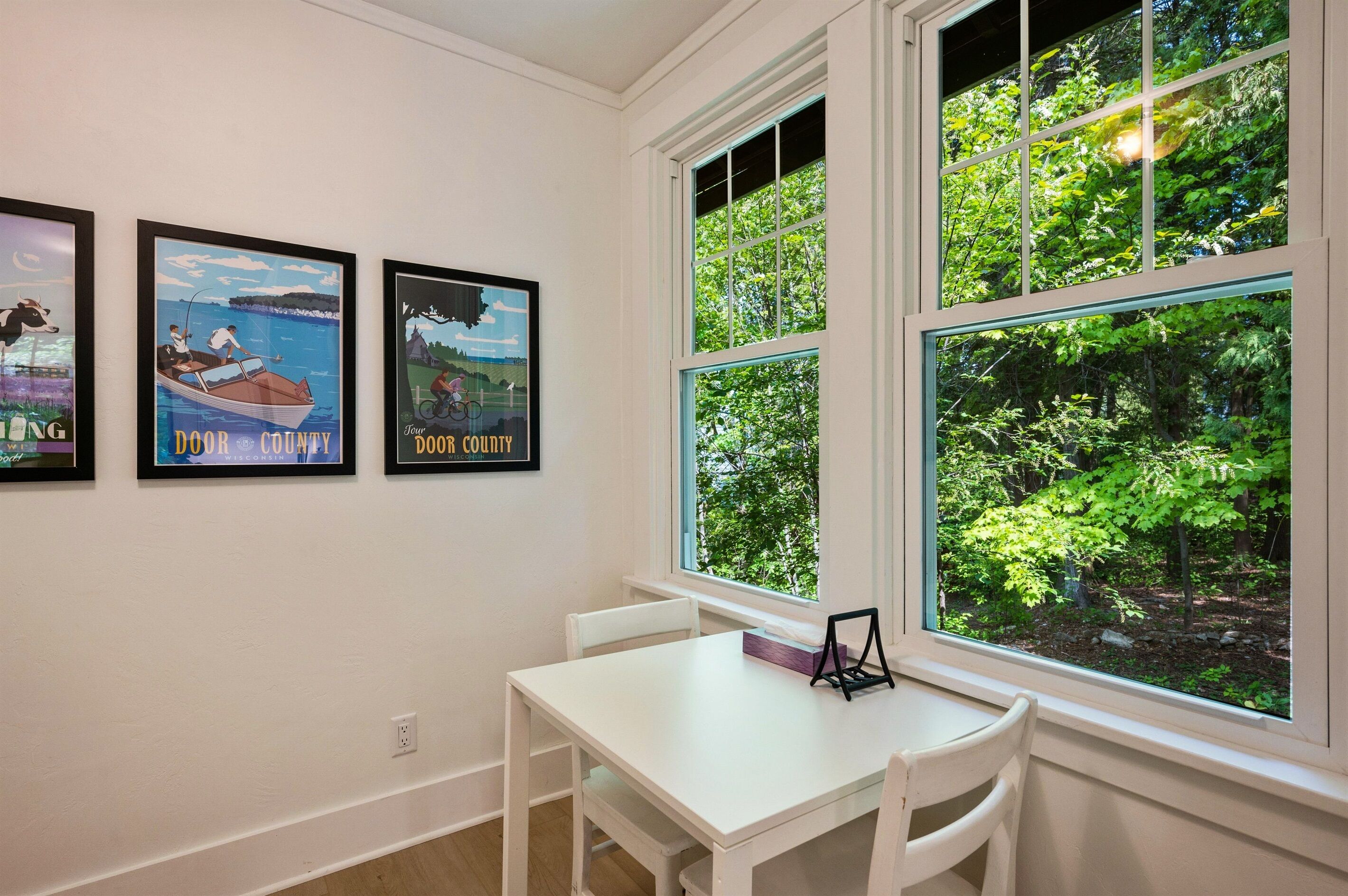 A room with a white table and chairs, a large window with a view of trees, and three framed pictures on the wall.