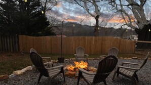 Un patio con quattro poltrone Adirondack disposte attorno a un braciere, sullo sfondo un recinto in legno, alberi e un tramonto.