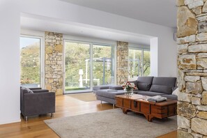 A modern living room with a stone accent wall, a grey sofa, a wooden coffee table, and large windows offering a view of greenery.
