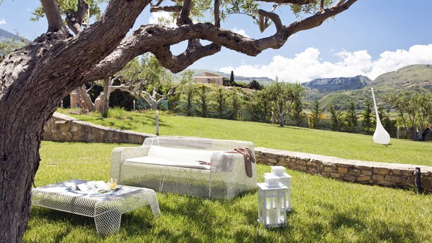 Ein Sitzbereich im Freien mit einem Tisch aus weißem Netzmaterial und einer weißen Couch unter einem Baum, mit einer Steinmauer und Blick auf eine hügelige Landschaft im Hintergrund.