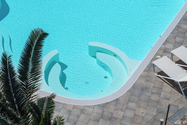 A swimming pool with a curved edge, surrounded by tiled flooring and palm trees.