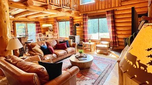 Un intérieur spacieux de chalet en bois, avec un grand plafond en bois, une cheminée, un coin salon confortable doté de canapés en cuir et un lustre.
