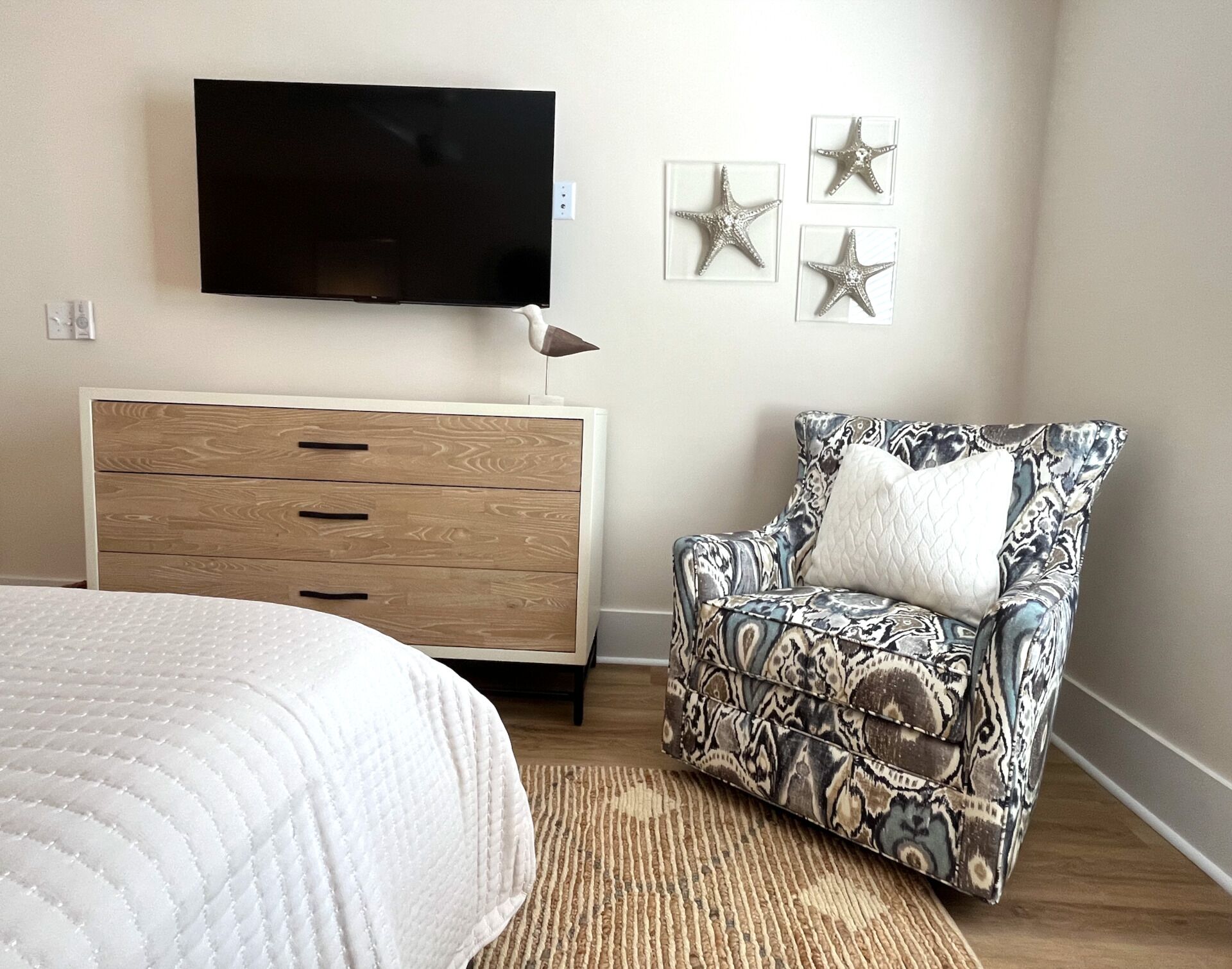 A bedroom with a bed, a wooden dresser, a patterned armchair, a television on the wall, and three star-shaped wall decorations.