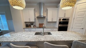 Una cuina moderna amb armaris blancs, tauleig de granit, electrodomèstics d'acer inoxidable i cadires de vimet.