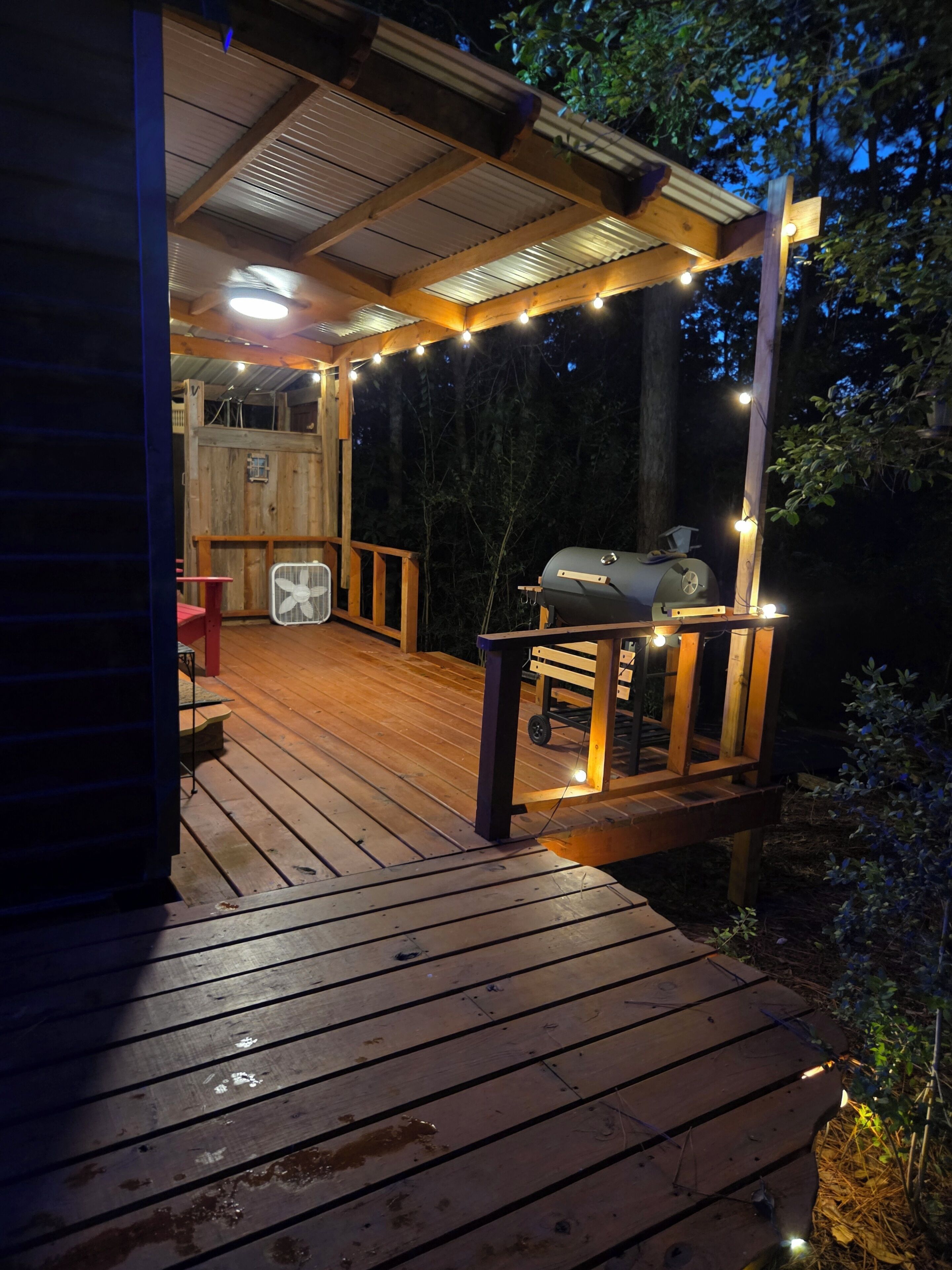 Une terrasse en bois avec un barbecue et des guirlandes lumineuses, entourée d’arbres.
