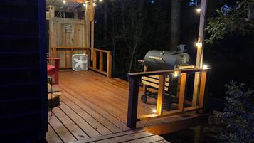 Une terrasse en bois avec un barbecue et des guirlandes lumineuses, entourée d’arbres.