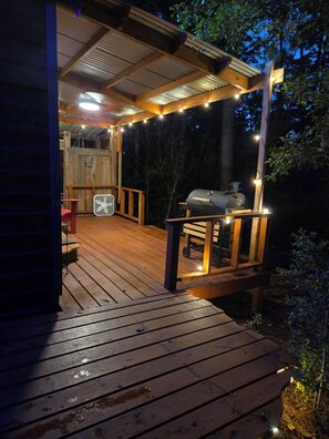 Une terrasse en bois avec un barbecue et des guirlandes lumineuses, entourée d’arbres.