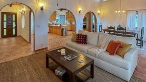 A spacious living area with a high ceiling, wooden beams, a ceiling fan, a sofa, a coffee table, and a kitchen in the background.