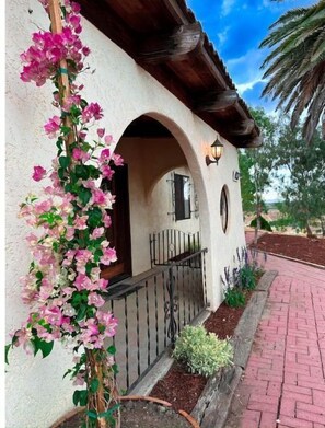 Un edificio con facciata in stucco, un balcone in legno e un giardino con un’edera fiorita di colore rosa.
