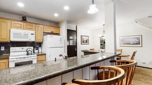 A modern kitchen with granite countertops, wooden cabinets, a microwave, stove, refrigerator, and bar stools. - The Summit 66 Walk to Eagle Lodge Chair 15 (Mammoth Lakes)