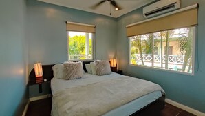 Une chambre à coucher comprenant un lit, un ventilateur de plafond, un appareil de climatisation, deux lampes de chevet et des fenêtres donnant sur un paysage verdoyant.