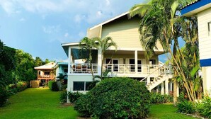 Une maison à deux étages dotée d’un balcon, entourée d’une végétation luxuriante et de palmiers.
