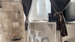 A room with a stone wall, a window with curtains, a desk with a computer, two white chairs, a small shelf with a lamp, and a floor vent.