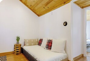 Une chambre avec un plafond en bois, un lit avec une literie blanche et des coussins à carreaux, un petit tabouret en bois sur lequel est posée une plante, et un mur blanc.