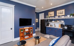 Et moderne køkken med mørkeblå skabe, et sort køleskab og en mikroovn. Der er en træbogreol med bøger og dekorative genstande, samt et fladskærms-TV monteret på væggen.