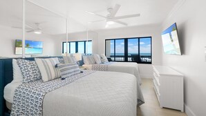 A bedroom with two beds, a ceiling fan, a TV, and a view of the ocean.