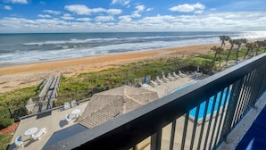 A balcony with a view of a beach, a pool, and lounge chairs.