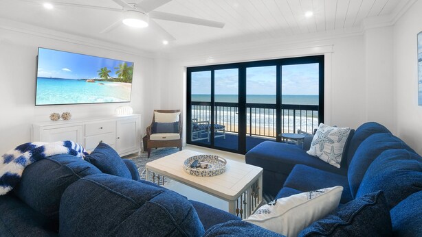A living room with a large TV, a ceiling fan, a sofa, a coffee table, and a view of the beach through large windows.