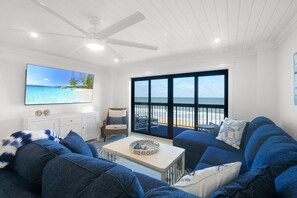 A living room with a large TV, a ceiling fan, a sofa, a coffee table, and a view of the beach through large windows. - Luxury direct oceanfront 3/3 corner with large private balcony, heated pool and garage (Ormond Beach)