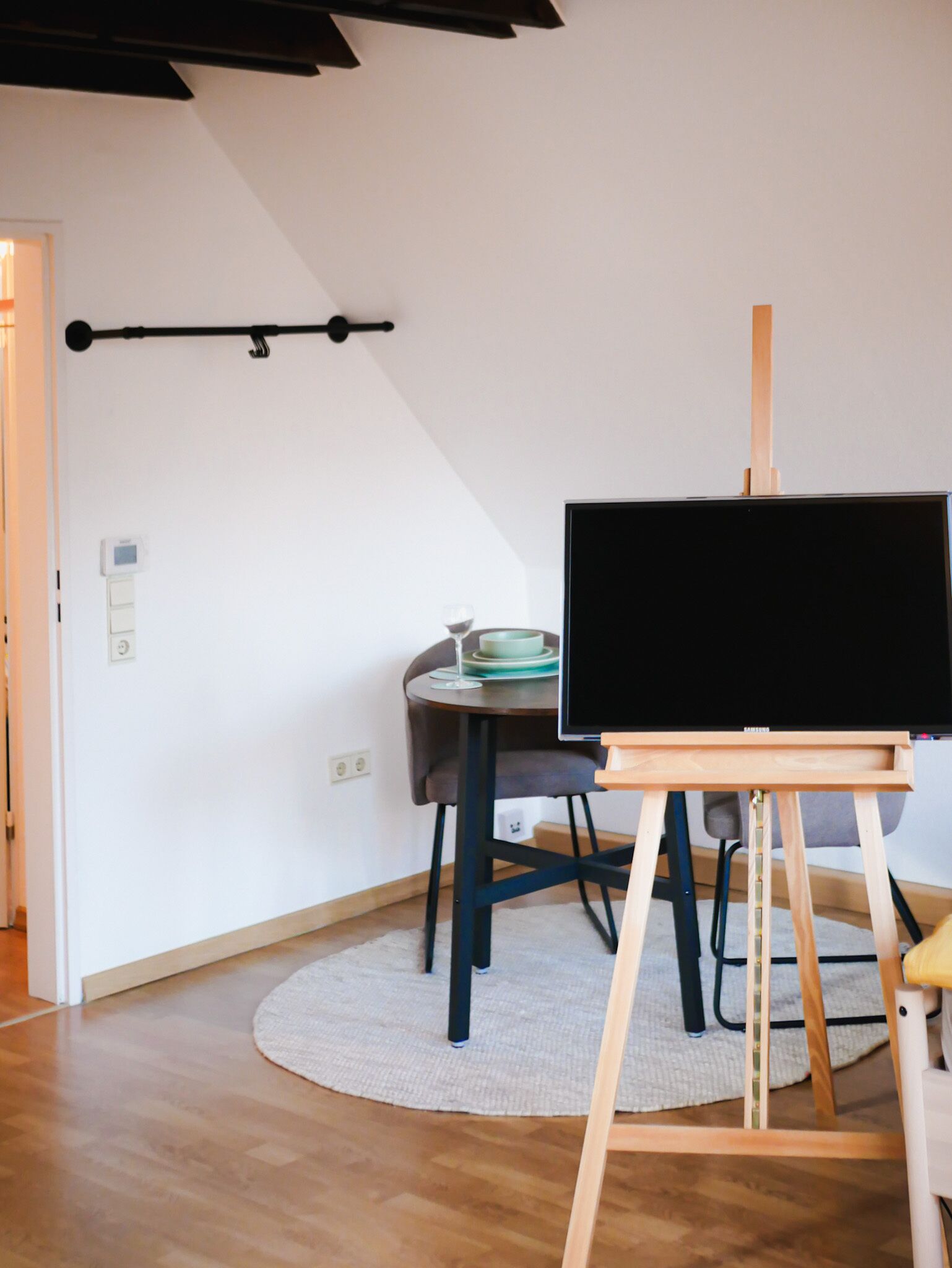 Een kamer met een houten vloer, een rond vloerkleed, een eettafel met stoelen, een televisie op een houten plank en een wandlamp.