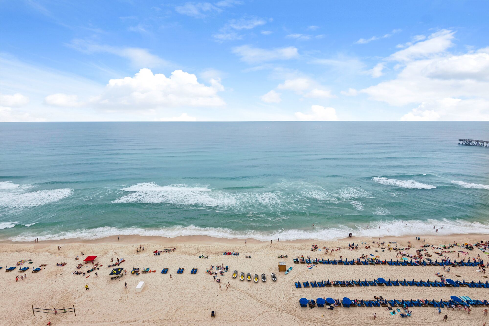 Ein Strand mit blauen Liegestühlen und Sonnenschirmen, einem Volleyballnetz und einem Pier in der Ferne.