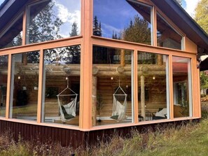Una cabaña con ventanas grandes, troncos de madera y dos hamacas.