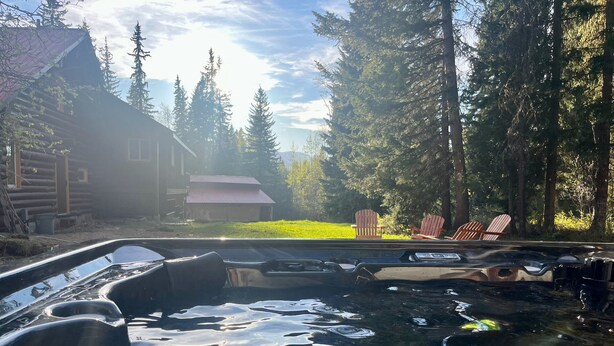 A hot tub surrounded by trees, a wooden cabin, and outdoor chairs. - Family Cabin + Games Barn | Hot Tub | Theatre (Columbia-Shuswap)