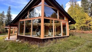 A log cabin with large windows, a wooden exterior, and a grassy surrounding. - Family Cabin + Games Barn | Hot Tub | Theatre (Columbia-Shuswap)