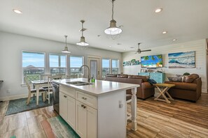 A spacious kitchen with a central island, a dining area with a table and chairs, a living room with a sofa and a coffee table, and a view of the ocean through large windows. - Beach Getaway. 2 King Beds. Walk to Beach. Fire Pit, BBQ Grill and Ping Pong (Galveston)
