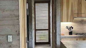 Een smalle, industriële hal met ruwe betonnen muren, een houten deur met horizontale zonwering, en een keuken met houten keukenkastjes en een gasfornuis.