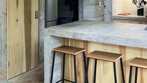 Een moderne keuken met een betonnen plafond, houten kastjes, een roestvrijstalen koelkast, een zwarte hanglamp en een bar met krukken.