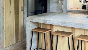Una cocina moderna con techo de concreto, armarios de madera, refrigerador de acero inoxidable, una lámpara colgante negra y una zona de bar con taburetes.