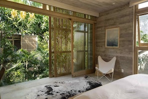 A bedroom with a large sliding glass door leading to a lush garden, a white bed, and a white chair.