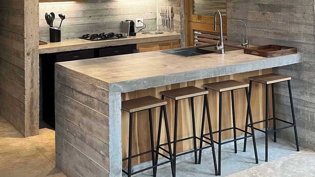 A modern kitchen with a large island, bar stools, wooden cabinets, and a window with blinds. - Casa Macu - Surf Oasis: Full House w/ Pools Steps From Waves (Playa La Saladita)