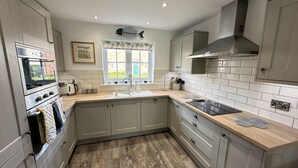 A modern kitchen with grey cabinets, a built-in oven, a sink, and a window with a view of greenery. - Stunning Coastal Cottage - Sleeping 6 in 3 Bedrooms - Pool and Beach Access Included. (Filey)
